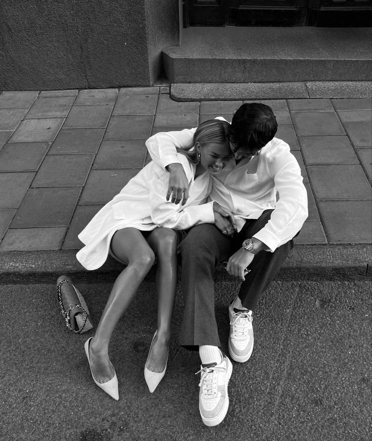 black and white photograph of two people sitting next to each other on the ground with their arms around each other