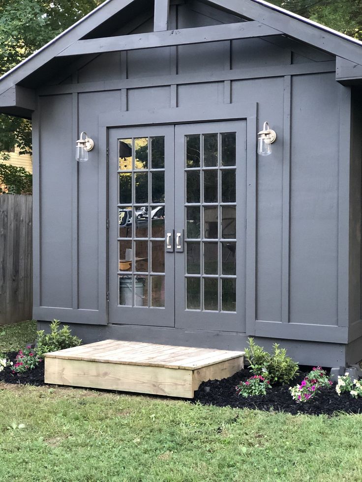 a small gray shed sitting in the grass next to a wooden bench and flower bed