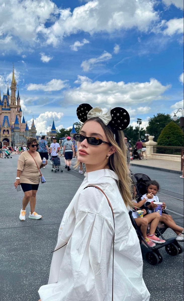 a woman wearing minnie mouse ears standing in front of a castle with people walking around