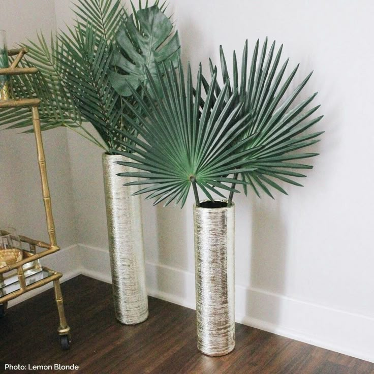 two metal vases with plants in them on a wooden floor next to a wall