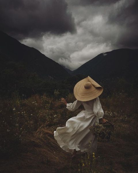 a woman in a white dress and straw hat walking through a field with mountains in the background