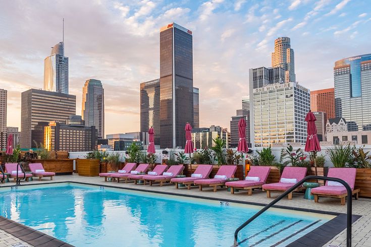 an outdoor swimming pool with pink lounge chairs and city skyline in the background at sunset