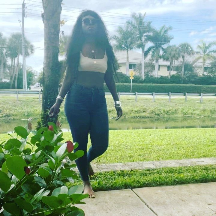 a woman walking down a sidewalk next to a lush green park with palm trees in the background