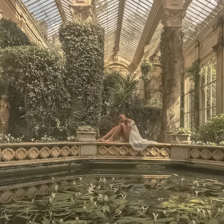 a woman sitting on the edge of a pond in front of a glass ceilinged building