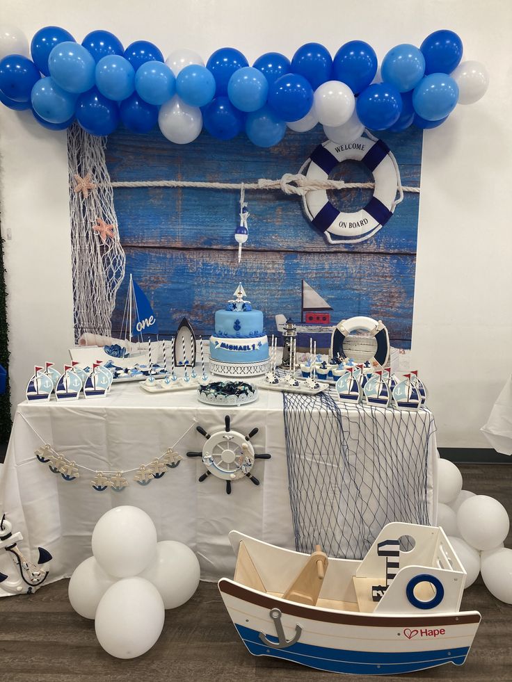 a table topped with blue and white balloons next to a boat filled with cake on top of it