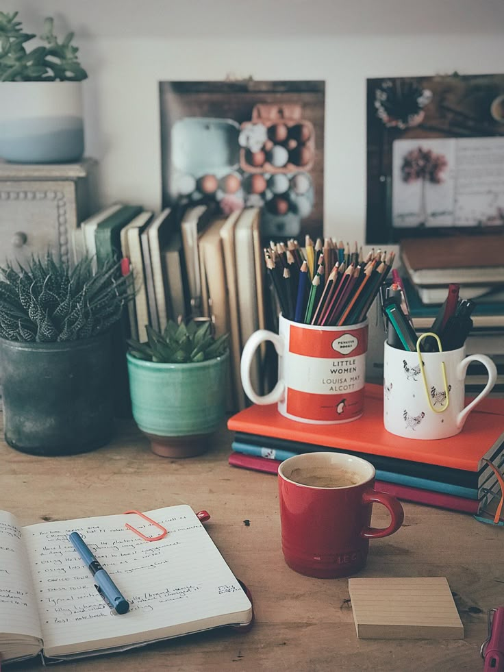 a desk with books, cups and pencils on it next to an open notebook