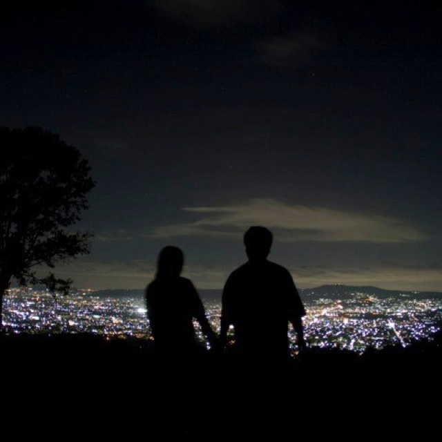 two people standing on top of a hill looking at the city lights in the distance