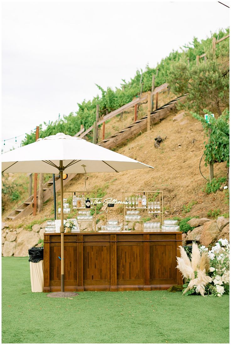 an outdoor bar set up with white umbrellas and flowers on the grass in front of a hill