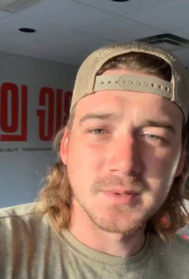 a man with long hair wearing a hat and looking at the camera while standing in front of a sign