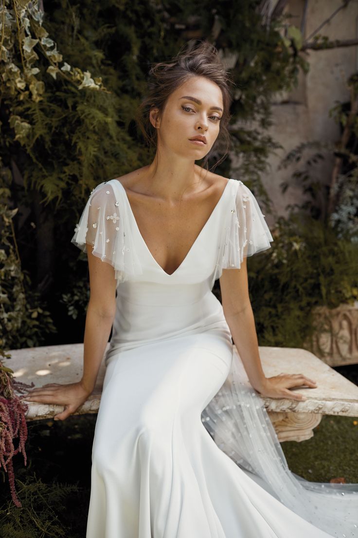 a woman is sitting on a bench wearing a white wedding dress with sheer sleeves and beading