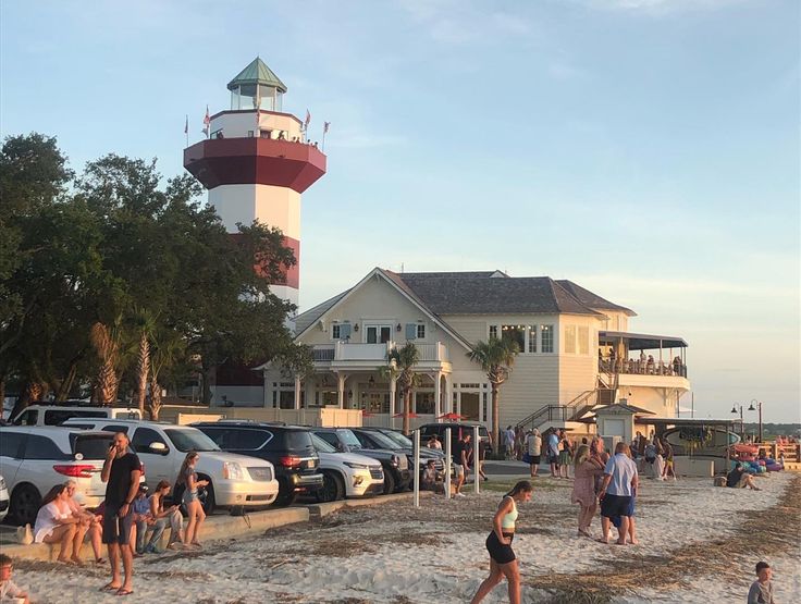 many people are walking on the beach near some cars and a lighthouse in the background