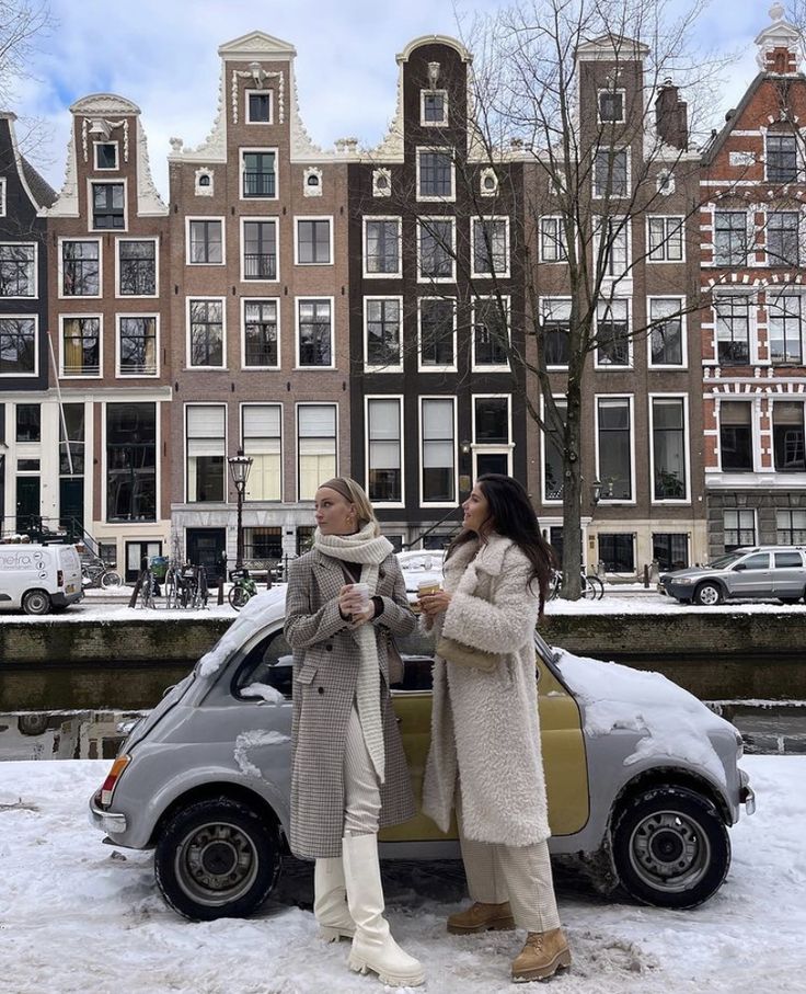 two women standing next to a small car in front of some buildings with snow on the ground