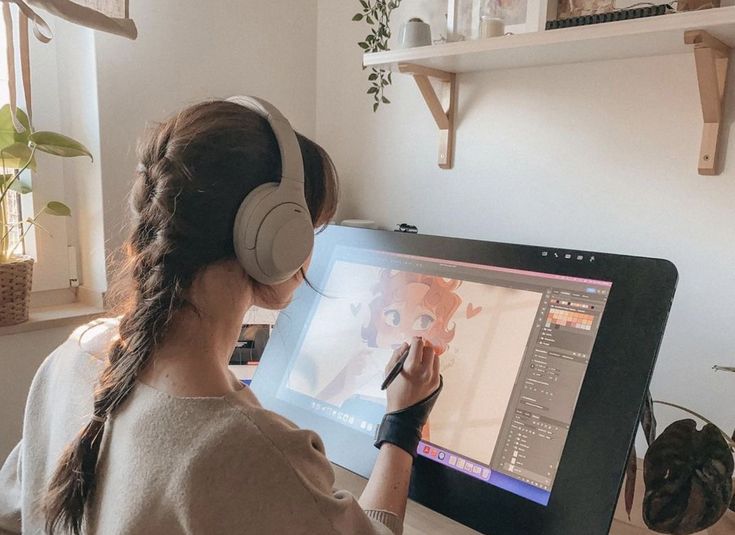 a woman sitting at a desk with headphones on, using a computer screen to draw pictures