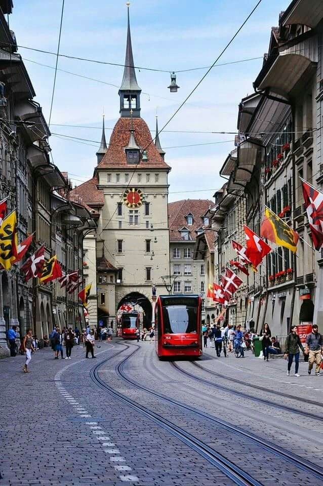 a red train is coming down the tracks in an old european city with people walking around