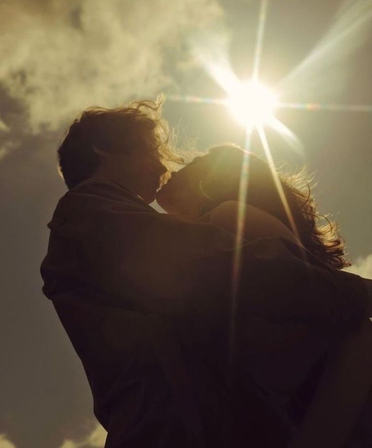 a man and woman kissing in front of the sun on a clear day with clouds