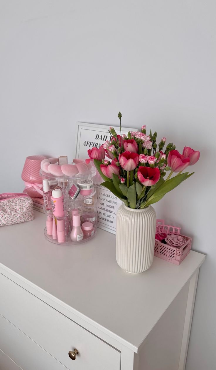 a white vase filled with pink flowers sitting on top of a dresser next to other items