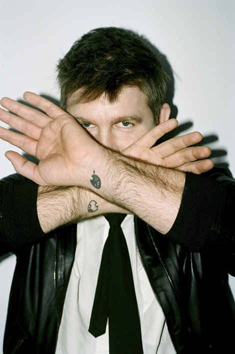 a man wearing a black leather jacket and tie covering his face with both hands while standing in front of a white wall