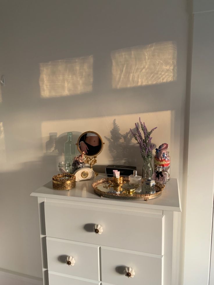 a white dresser topped with lots of drawers next to a mirror and vase filled with flowers