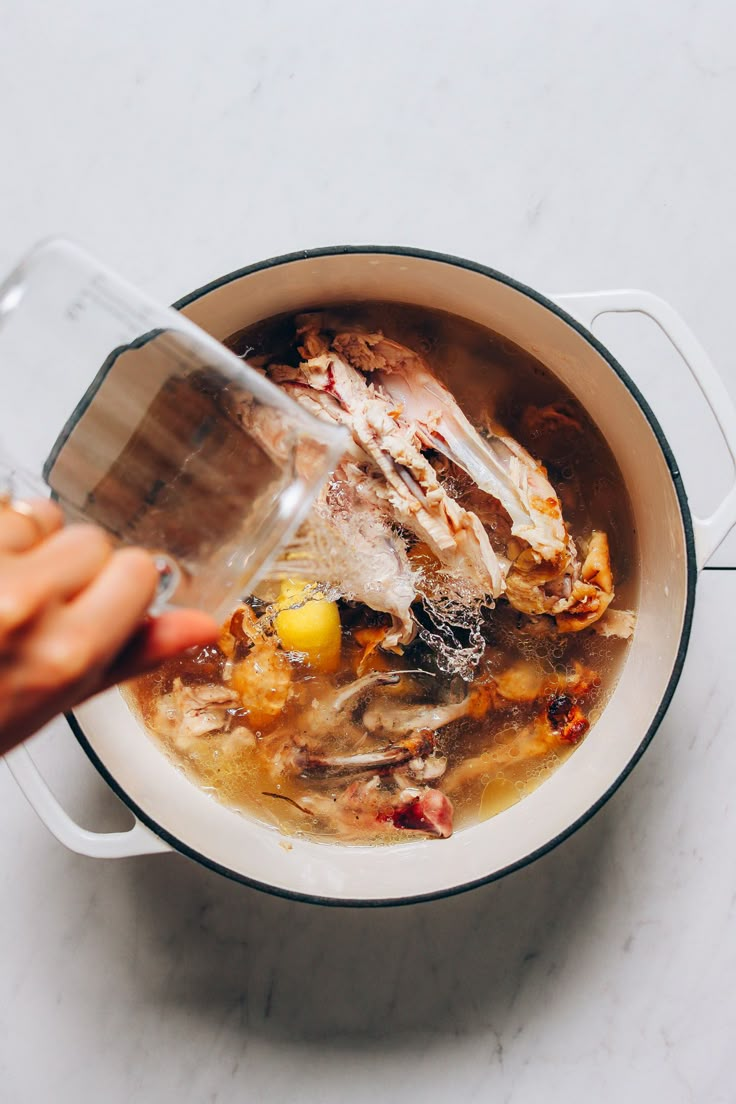 a person pouring water into a pot filled with chicken and vegetable soup on top of a white counter