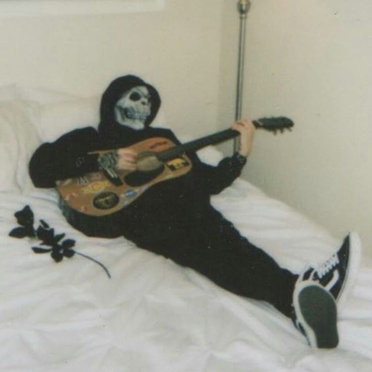 a person in a skeleton mask playing an acoustic guitar on a bed with white sheets