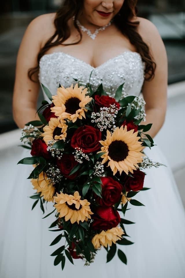 a bride holding a bouquet of sunflowers and red roses in her wedding dress