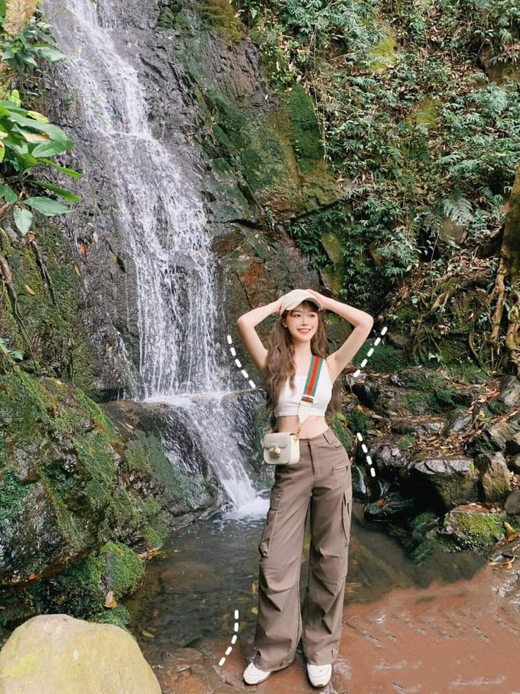 a woman standing in front of a waterfall with her hands on her head and looking at the camera