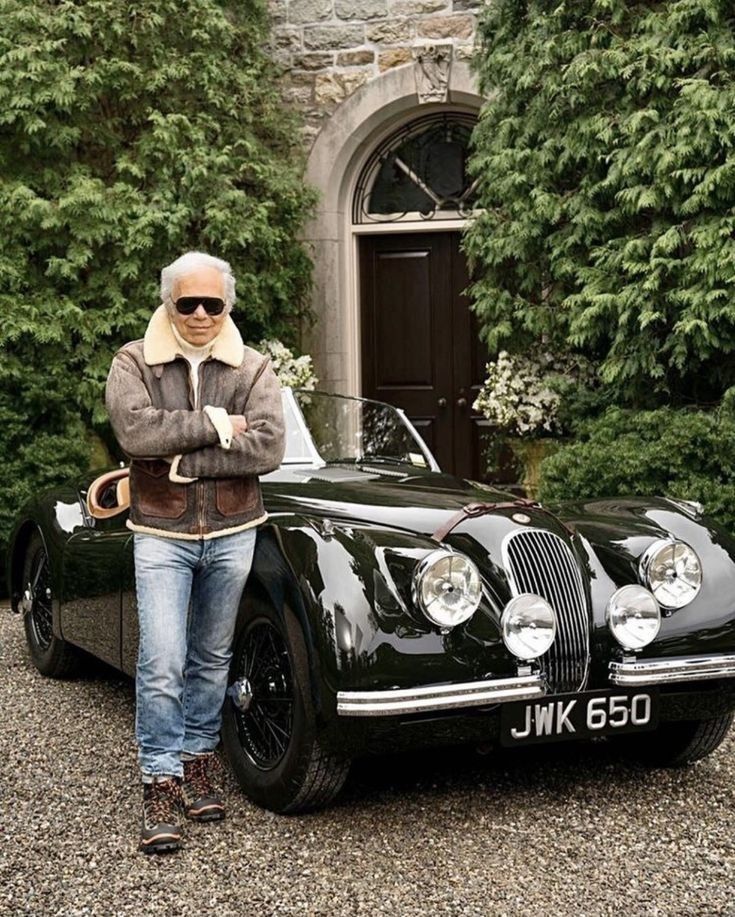 an older man standing next to a black car in front of a stone building with trees