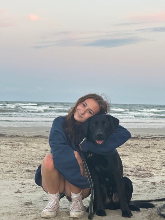 a woman kneeling down next to a black dog on top of a sandy beach with the ocean in the background