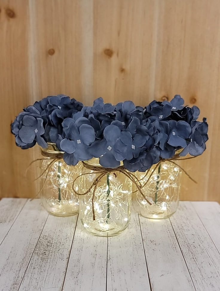 three glass vases filled with blue flowers on top of a white wooden table next to a wood wall