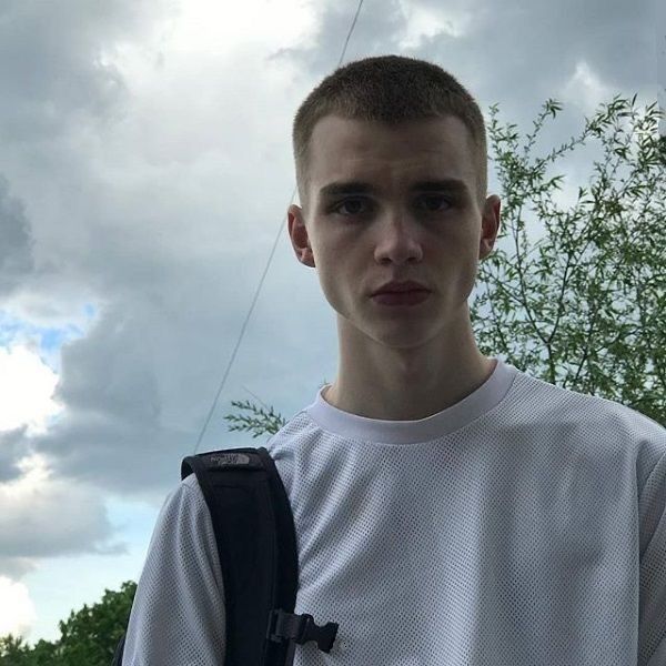 a young man with a back pack on his shoulder standing in front of some trees