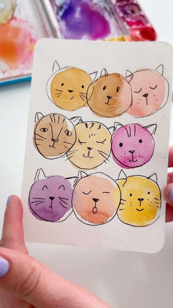 a person holding up a piece of paper with cats drawn on it and watercolor paints in the background