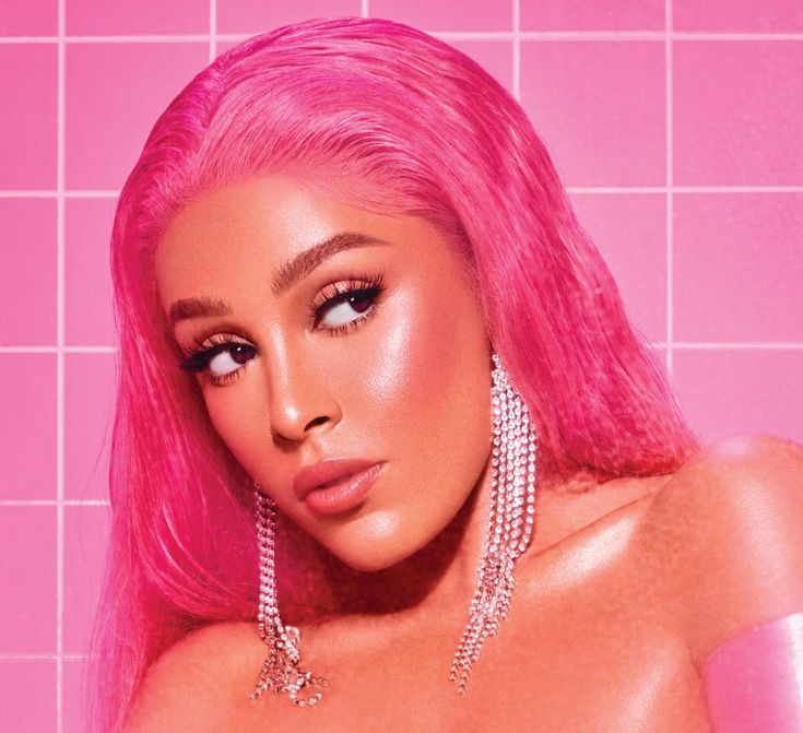 a woman with pink hair and earrings in front of a tiled wall, looking at the camera