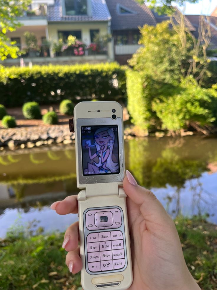 a person holding up a cell phone in front of a pond and house with trees