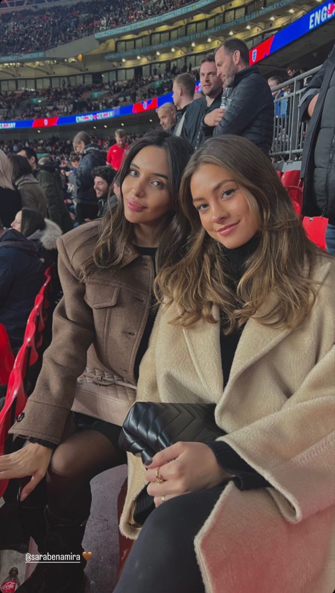 two women sitting next to each other at a soccer game in the stands, one wearing a tan coat