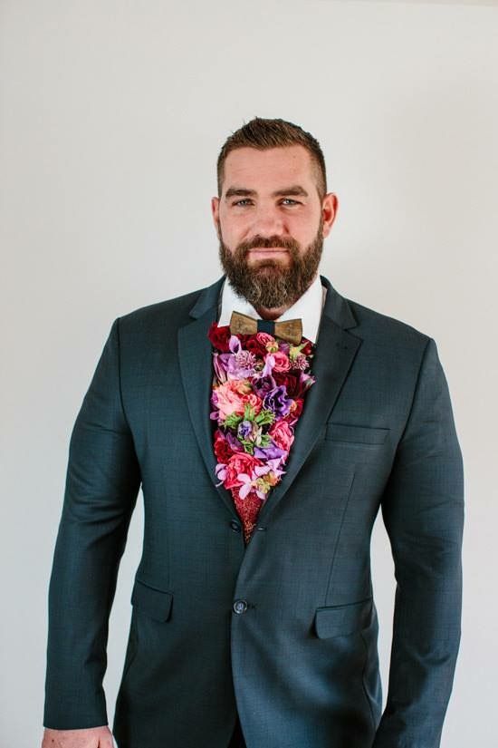 a man with a beard wearing a suit and flowered bow tie standing in front of a white wall