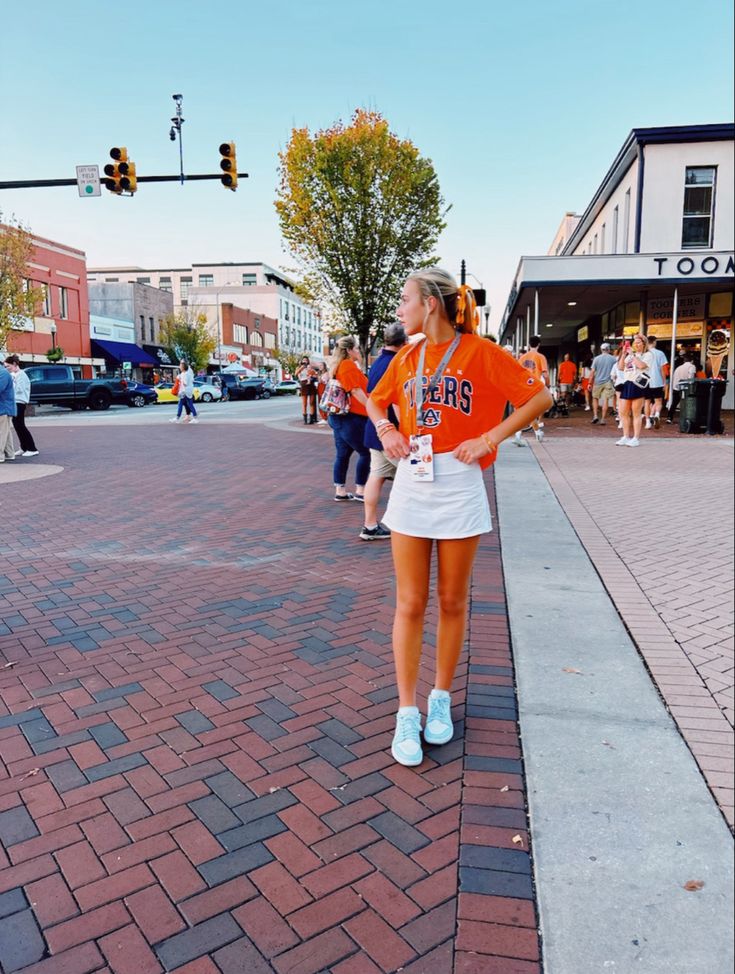 a woman in an orange shirt and white skirt standing on the sidewalk with her hands behind her back