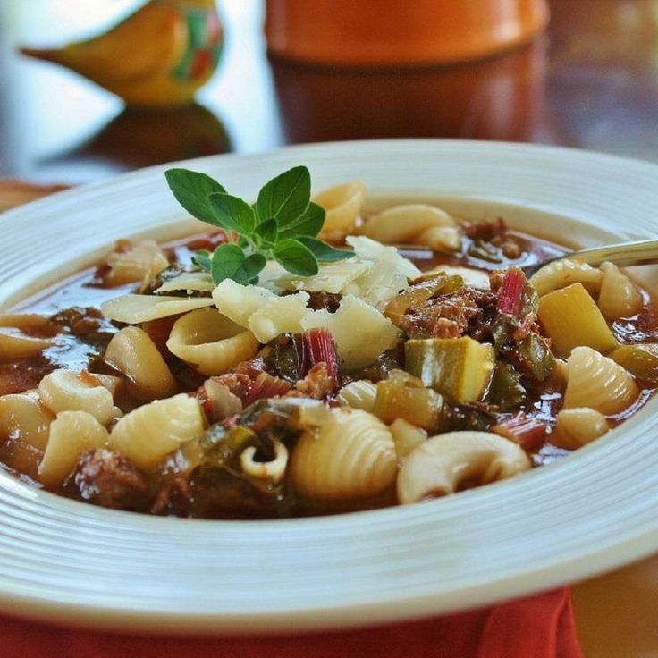 a white bowl filled with pasta and meat soup on top of a red cloth covered table