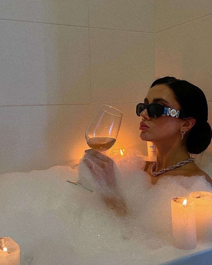 a woman sitting in a bathtub holding a glass of wine with candles around her