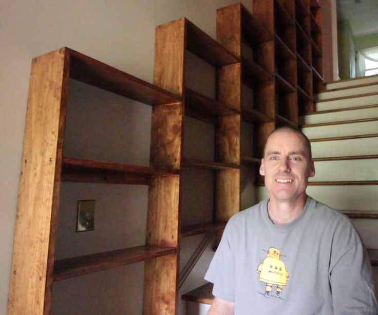 a man standing in front of a wooden book shelf with shelves on each side and stairs up to the second floor