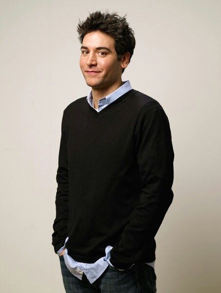 a man standing in front of a white wall wearing a black sweater and blue shirt