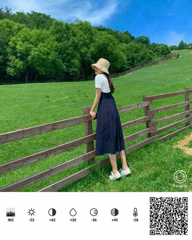 a woman leaning on a fence looking at the grass and trees in the field behind her