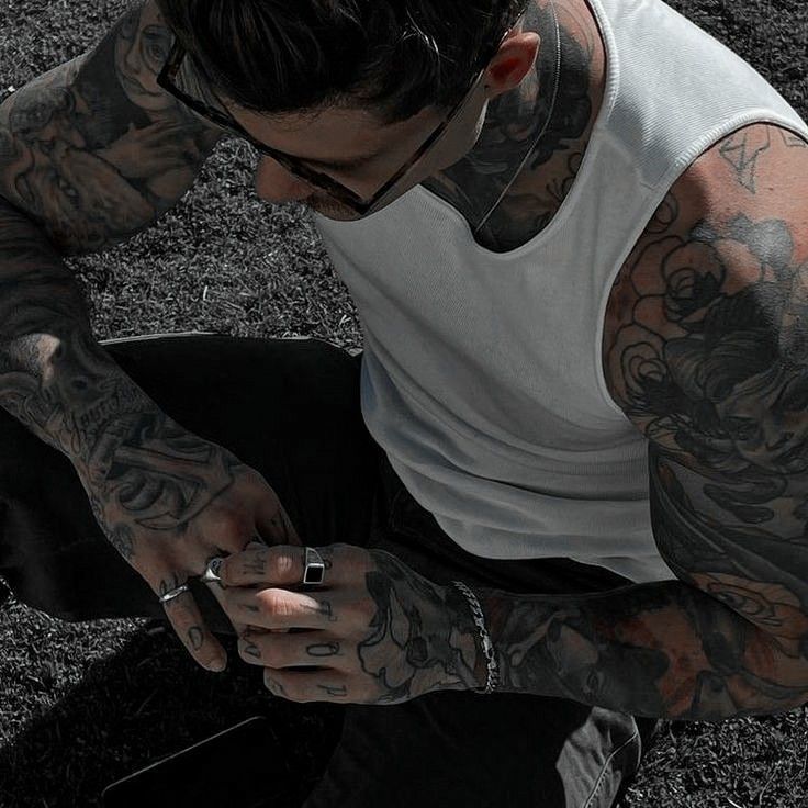 a man sitting in the grass with tattoos on his arms and legs, looking at his cell phone