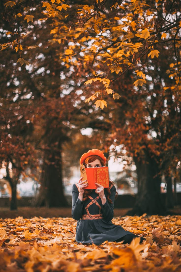 a woman is sitting in the leaves with an orange hat on her head and she is holding up a book