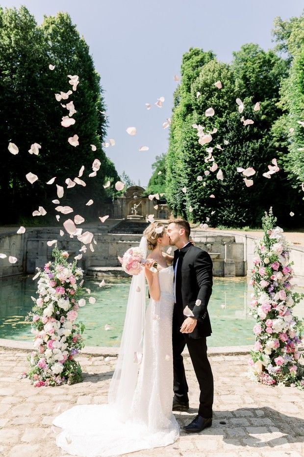 a newly married couple kissing in front of a fountain with pink flowers and petals falling from it