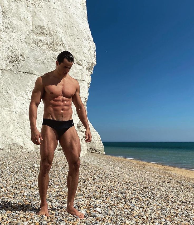 a man standing on top of a rocky beach next to the ocean with his bare chest