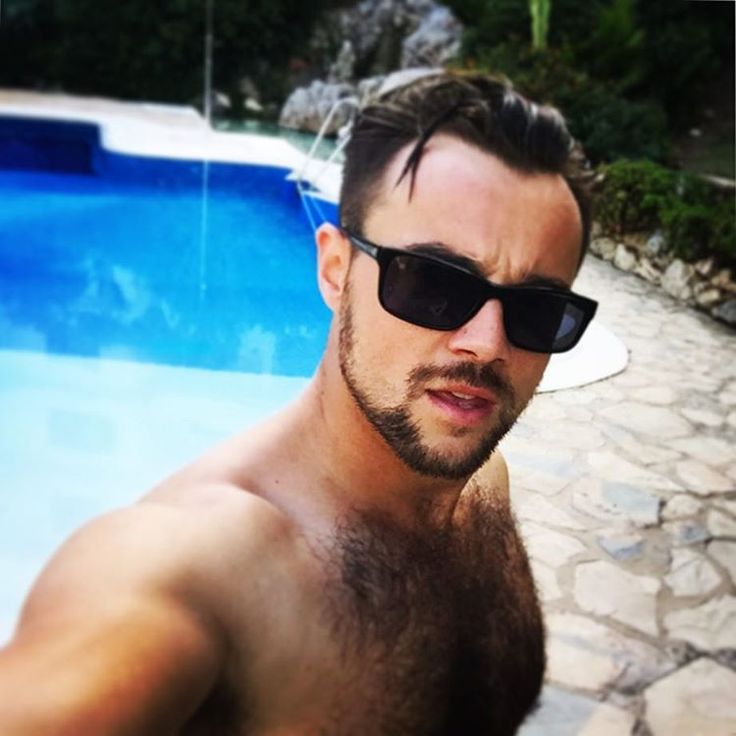 a shirtless man wearing sunglasses standing in front of a swimming pool and looking at the camera