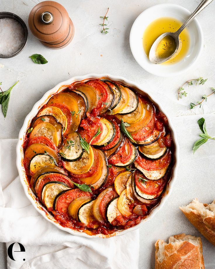 a dish filled with sliced vegetables next to bread and olive oil on a white table