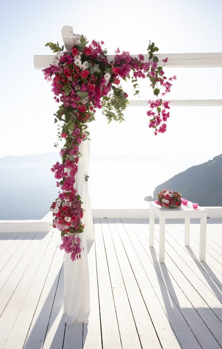 a wedding arch decorated with pink flowers and greenery on a deck overlooking the ocean