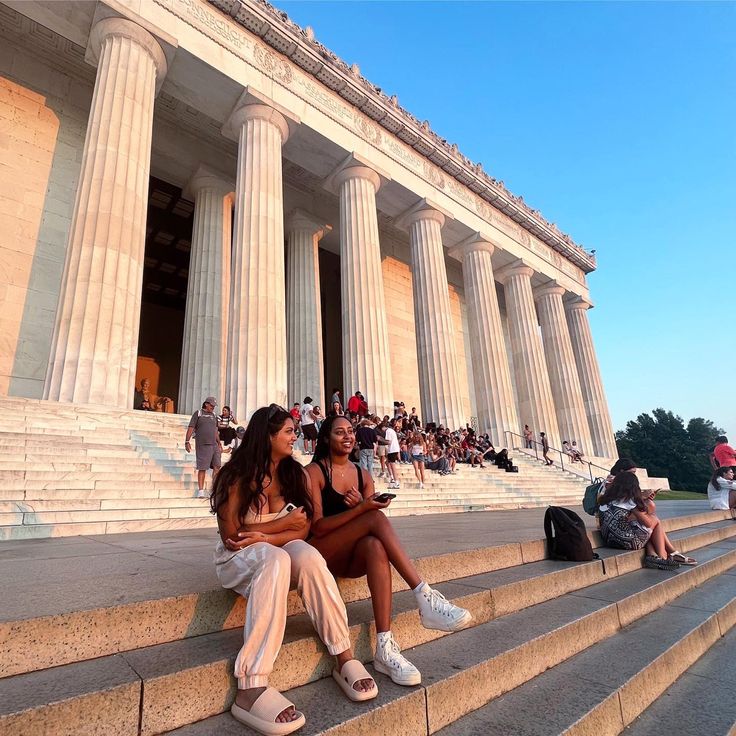 two women sitting on steps in front of a building with columns and people walking around