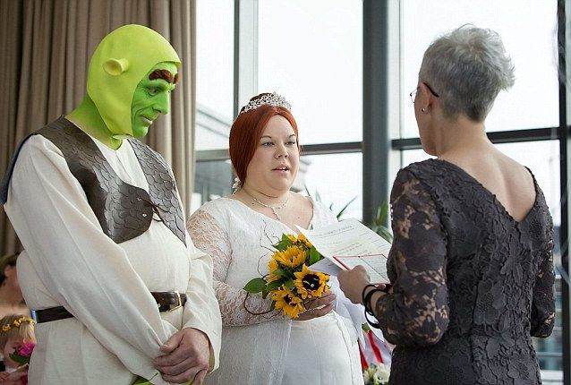 a woman in a white dress standing next to a man wearing a green mask and holding flowers